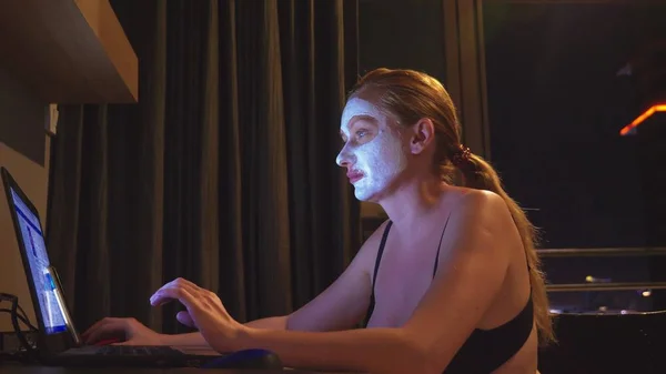 young woman with cosmetic mask on face working with laptop at home in the evening. naturalness
