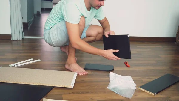 Um jovem coleta independentemente móveis na sala de estar de sua casa. Um homem recolhe uma secretária de computador . — Fotografia de Stock