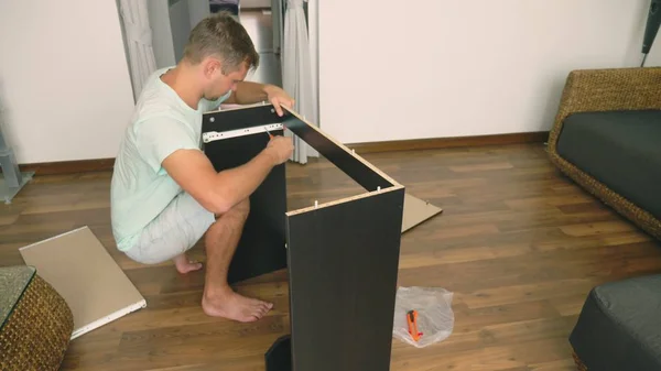 Een jonge man verzamelt zelfstandig meubels in de woonkamer van zijn huis. Een man verzamelt een computerbureau. — Stockfoto