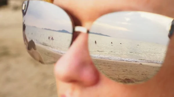 Közeli arcát egy boldog nő a napszemüveg. A strand, a nyugalmi emberek tükröződik a szemüveg. — Stock Fotó