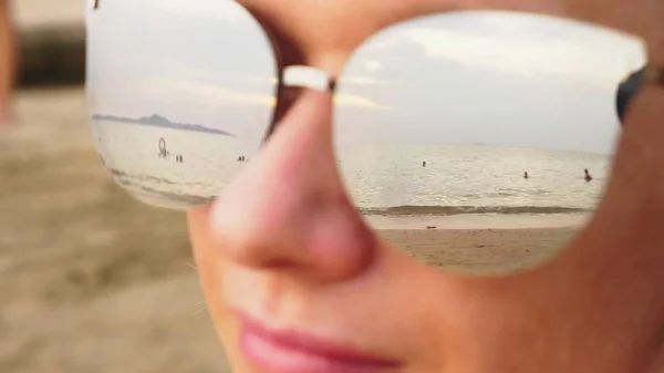 Wajah dekat dari wanita bahagia berkacamata hitam. Pantai dengan orang-orang yang beristirahat tercermin dalam kacamata . — Stok Foto