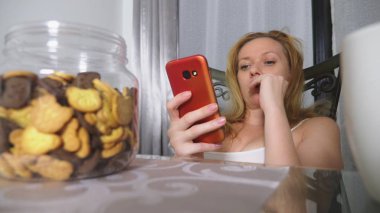 hungry sad woman sits in a living room at a table at night, she eats a liver and uses her smartphone clipart