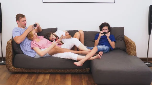 Charming family, mom, dad, daughter and son are watching TV in the living room together, everyone is looking into his phone — Stock Photo, Image