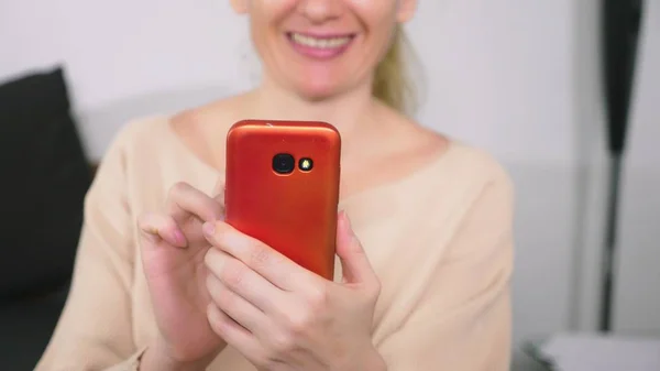 Bella ragazza utilizzare uno smartphone cellulare mentre seduto in soggiorno la sera. Felice sorridente giovane donna che chiacchiera su internet a casa . — Foto Stock