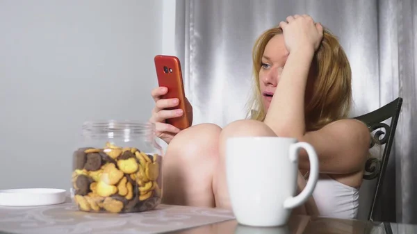 Hongerige triest vrouw zit in een woonkamer aan een tafel in de nacht, ze eet een lever en gebruikt haar smartphone — Stockfoto