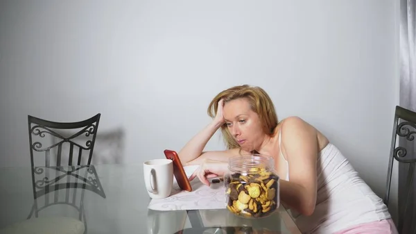 Hongerige triest vrouw zit in een woonkamer aan een tafel in de nacht, ze eet een lever en gebruikt haar smartphone — Stockfoto