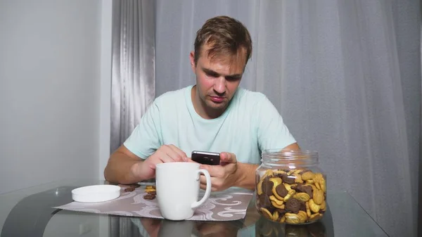 Hombre triste hambriento se sienta en una sala de estar en una mesa por la noche, se come un hígado y utiliza su teléfono inteligente —  Fotos de Stock