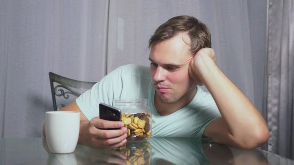 Hombre triste hambriento se sienta en una sala de estar en una mesa por la noche, se come un hígado y utiliza su teléfono inteligente — Foto de Stock