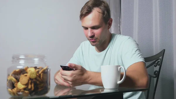 Hungry sad man sits in a living room at a table at night, he eats a liver and uses her smartphone — Stock Photo, Image