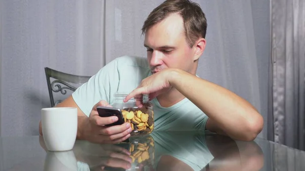 Hongerige trieste man zit in een woonkamer aan een tafel bij nacht, hij eet een lever en gebruikt haar smartphone — Stockfoto