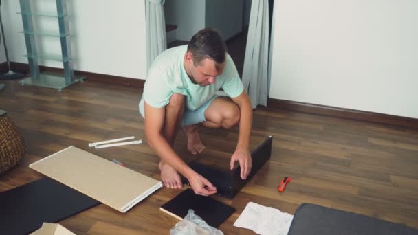 Un joven colecciona muebles de forma independiente en la sala de estar de su casa. Un hombre recoge un escritorio de computadora . — Vídeos de Stock