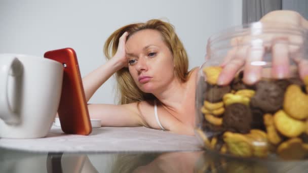 饥饿悲伤的女人晚上坐在客厅的桌子旁 她吃肝脏 用她的智能手机 — 图库视频影像