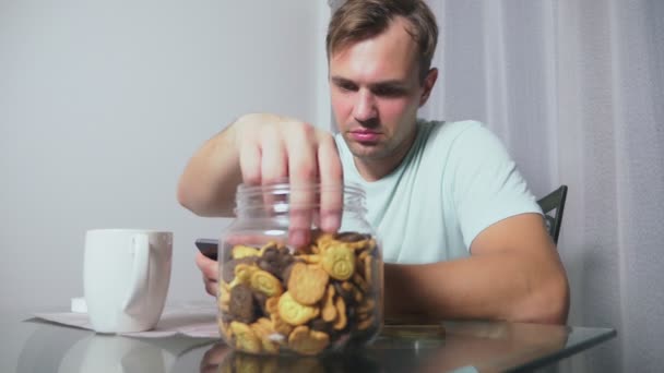 Hungrig ledsen man sitter i ett vardagsrum vid ett bord på natten, han äter en lever och använder sin smartphone — Stockvideo