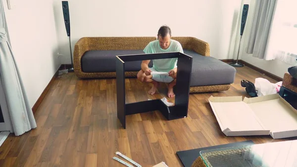 Een jonge man verzamelt zelfstandig meubels in de woonkamer van zijn huis. Een man verzamelt een computerbureau. — Stockfoto