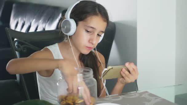 Teknoloji ve rekreasyon kavramı. Kulaklık içindeki herhangi bir dinleme ve telefon tarama evde rahatlatıcı, yemek salonunda masada oturan olumlu neşeli genç kız. yemek — Stok video