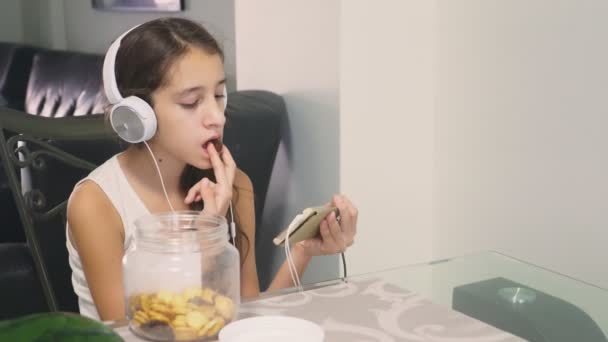 Het concept van technologie en recreatie. Positieve vrolijke tienermeisje ontspannen thuis, zitten aan de tafel in de eetkamer, luisteren naar iets in de hoofdtelefoon en de telefoon surfen. eten — Stockvideo