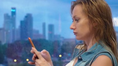 ıslak sarışın kız dışarıda yağmur yağdığında penceredeki gökdelenler çerçevede ayakta, ona telefon kullanır