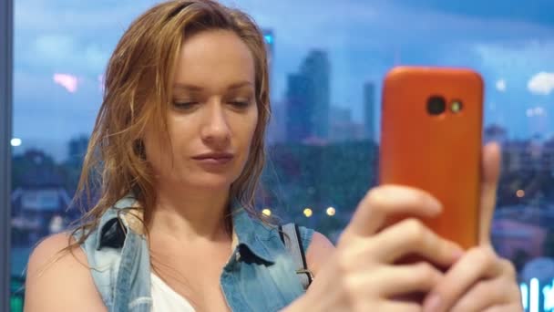 Nasses blondes Mädchen, das vor dem Hintergrund von Wolkenkratzern am Fenster steht, benutzt ihr Handy, während es vor dem Fenster regnet — Stockvideo