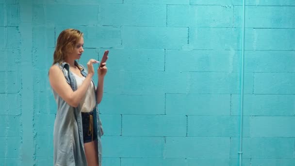 Menina loira atraente usa smartphone vermelho contra uma parede azul — Vídeo de Stock