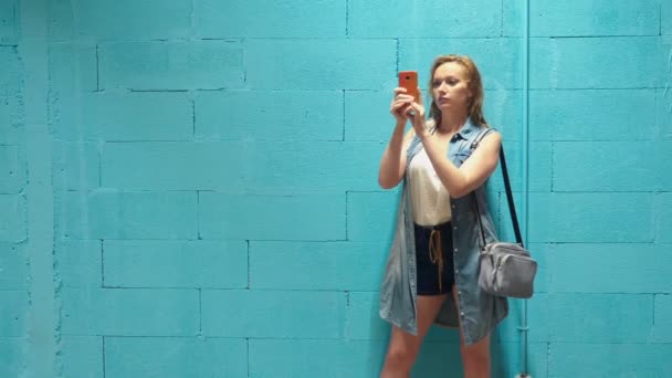 Attractive blonde girl uses red smartphone against a blue wall — Stock Video