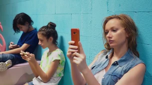 Garçon, fille et femme utilisent leur téléphone portable tout en étant assis contre le mur bleu — Video