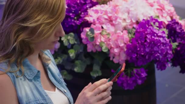 Egy gyönyörű fiatal nő ül egy padon egy nagy modern bevásárlóközpont közelében egy virágágyás. ő használ neki smartphone — Stock videók