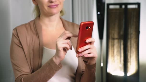 Beautiful girl use a cellular smartphone while sitting in the living room in the evening. Happy smiling young woman chatting on the internet at home. — Stock Video