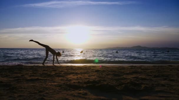 Σιλουέτα ενός κοριτσιού στο ηλιοβασίλεμα, με φόντο την θάλασσα, λεπτό κορίτσι μακροπόδαρος αστεία κάνουν γυμναστικές πραξικοπήματα στην ακτή της θάλασσας — Αρχείο Βίντεο