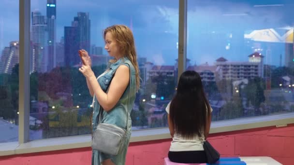 湿的金发碧眼的女孩站在摩天大楼的背景下, 在窗口, 用她的手机, 而窗外下雨 — 图库视频影像