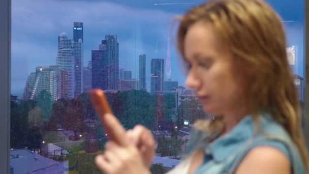 Nasses blondes Mädchen, das vor dem Hintergrund von Wolkenkratzern am Fenster steht, benutzt ihr Handy, während es vor dem Fenster regnet — Stockvideo