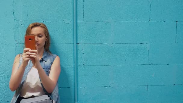 迷人的金发女孩使用红色智能手机对蓝色的墙壁 — 图库视频影像