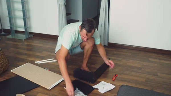 Een jonge man verzamelt zelfstandig meubels in de woonkamer van zijn huis. Een man verzamelt een computerbureau. — Stockfoto