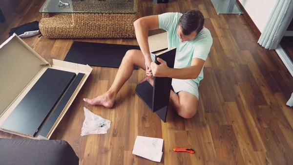 Um jovem coleta independentemente móveis na sala de estar de sua casa. Um homem recolhe uma secretária de computador . — Fotografia de Stock