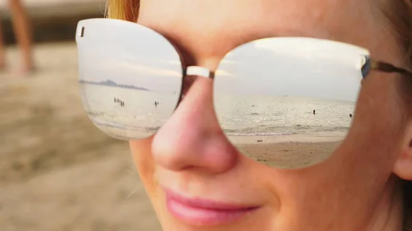ใบหน้าใกล้ชิดของผู้หญิงที่มีความสุขในแว่นกันแดด ชายหาดที่มีคนพักผ่อนสะท้อนในแว่นตา . — ภาพถ่ายสต็อก