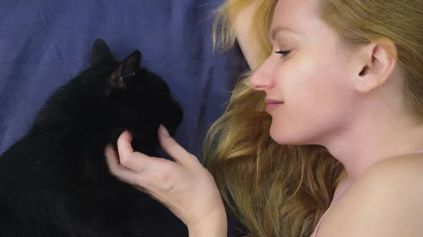 猫と少女 美しいブロンドの女の子のベッドに横たわっていると 彼女の黒い猫を Caressing します 動物の世話 ペットのための愛 — ストック写真