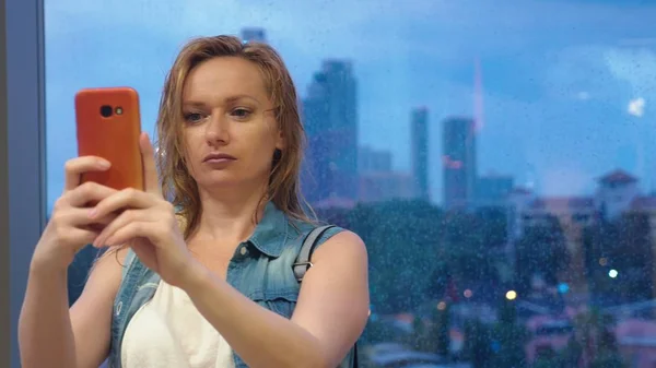 Nasses blondes Mädchen, das vor dem Hintergrund von Wolkenkratzern am Fenster steht, benutzt ihr Handy, während es vor dem Fenster regnet — Stockfoto