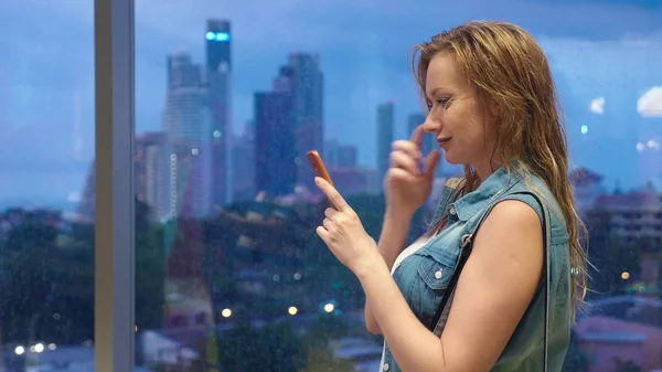 Nasses blondes Mädchen, das vor dem Hintergrund von Wolkenkratzern am Fenster steht, benutzt ihr Handy, während es vor dem Fenster regnet — Stockfoto