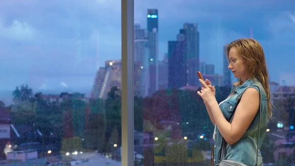 Mokré blonďaté dívky stojící u okna na pozadí mrakodrapů, používá její telefon, zatímco prší za oknem — Stock fotografie