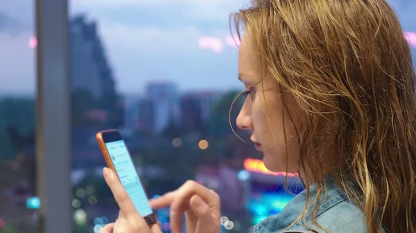 Nasses blondes Mädchen, das vor dem Hintergrund von Wolkenkratzern am Fenster steht, benutzt ihr Handy, während es vor dem Fenster regnet — Stockfoto