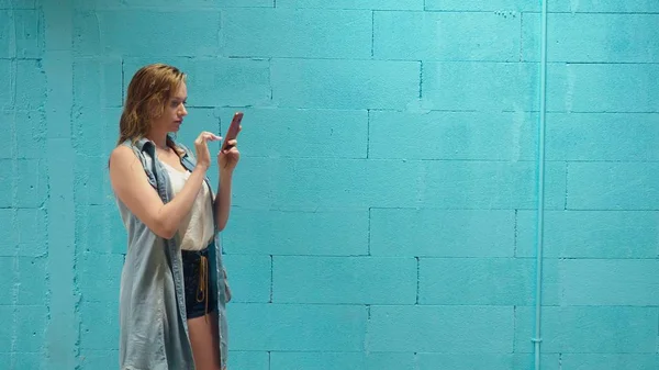 Gadis pirang yang menarik menggunakan smartphone merah menghadap dinding biru — Stok Foto