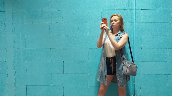 Attraktives blondes Mädchen nutzt rotes Smartphone gegen blaue Wand — Stockfoto