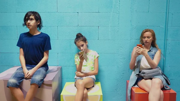 Niño, niña y mujer usan sus teléfonos móviles mientras están sentados contra la pared azul —  Fotos de Stock