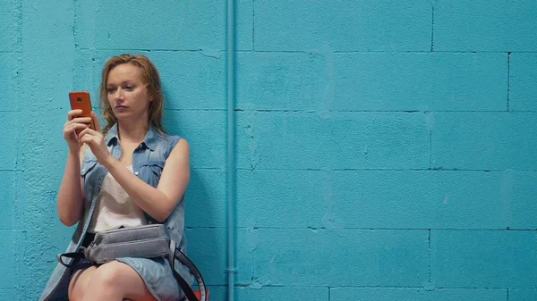 Atrakcyjna blondynka używa czerwony smartfona pod ścianą, niebieski — Zdjęcie stockowe