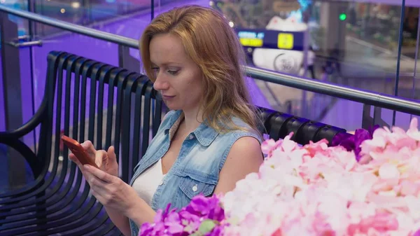 Eine schöne junge Frau sitzt auf einer Bank in einem großen modernen Einkaufszentrum in der Nähe eines Blumenbeets. Sie benutzt ihr Smartphone — Stockfoto