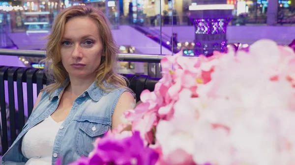 Uma bela jovem está sentada em um banco em um grande centro comercial moderno perto de um canteiro de flores . — Fotografia de Stock