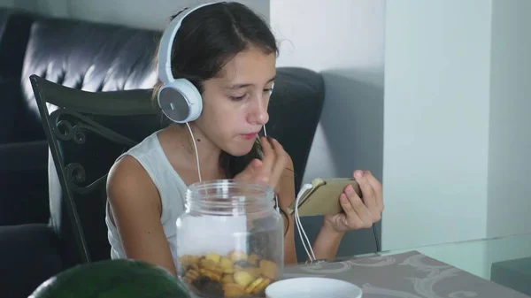 El concepto de tecnología y recreación. Chica adolescente alegre positivo relajarse en casa, sentado en la mesa en el comedor, escuchando algo en los auriculares y navegar por el teléfono. comer — Foto de Stock