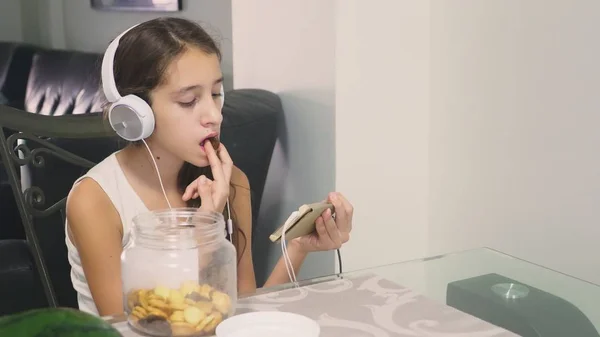 El concepto de tecnología y recreación. Chica adolescente alegre positivo relajarse en casa, sentado en la mesa en el comedor, escuchando algo en los auriculares y navegar por el teléfono. comer — Foto de Stock