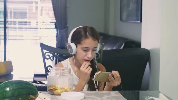 Konsep teknologi dan rekreasi. Gadis remaja yang gembira santai di rumah, duduk di meja di ruang makan, mendengarkan sesuatu dengan headphone dan meramban telepon. makan — Stok Foto