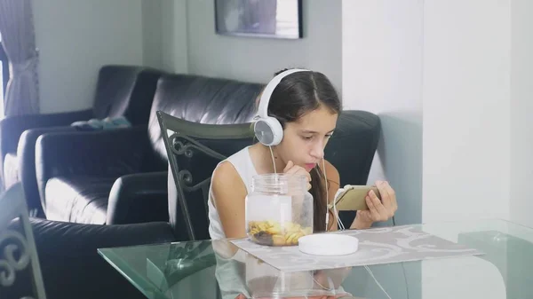 El concepto de tecnología y recreación. Chica adolescente alegre positivo relajarse en casa, sentado en la mesa en el comedor, escuchando algo en los auriculares y navegar por el teléfono. comer — Foto de Stock