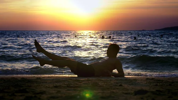 Chico guapo juguetón en pantalones cortos haciendo ejercicio, en la orilla del mar contra el telón de fondo de una maravillosa puesta de sol — Foto de Stock
